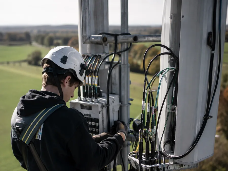 A telecommunications apprentice in a hard hat and harness at the base of a cell tower.